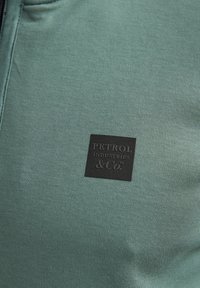 Tela de algodón verde con una textura suave y una etiqueta cuadrada negra que dice "PETROL INDUSTRIES & Co" se muestra de manera destacada.