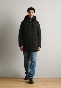 Parka negra con capucha, presenta dos bolsillos en el pecho, corte recto, llevada sobre un suéter gris, combinada con jeans de mezclilla y zapatos negros.