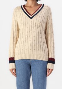 Beige kaapoknitti neule, jossa on syvä V-pääntie, tummansininen ja burgundinpunainen raidoitus kauluksessa ja hihansuissa, ribattu helma ja pitkät hihat.