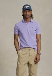 Mladik s dredovima nosi plavu kapu "Ralph", svijetlopurpuroljasti polo majicu, bež hlače, sat na ruci i stoji s rukama u džepovima.