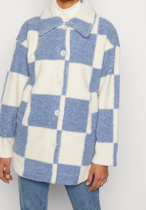 Veste en polaire à carreaux bleus et blancs boutonnée, portée par-dessus un col roulé blanc, associée à un jean bleu.