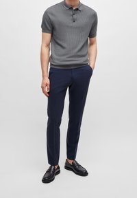 Homme portant un polo en maille à manches courtes gris, un pantalon tailleur bleu marine et des mocassins en cuir noirs, se tenant avec une main dans sa poche.