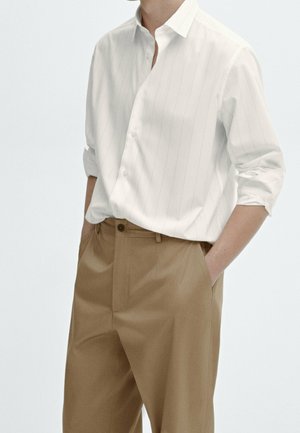 Personne portant une chemise blanche à fines rayures avec manches longues retroussées, rentrée dans un pantalon marron taille haute, mains dans les poches.