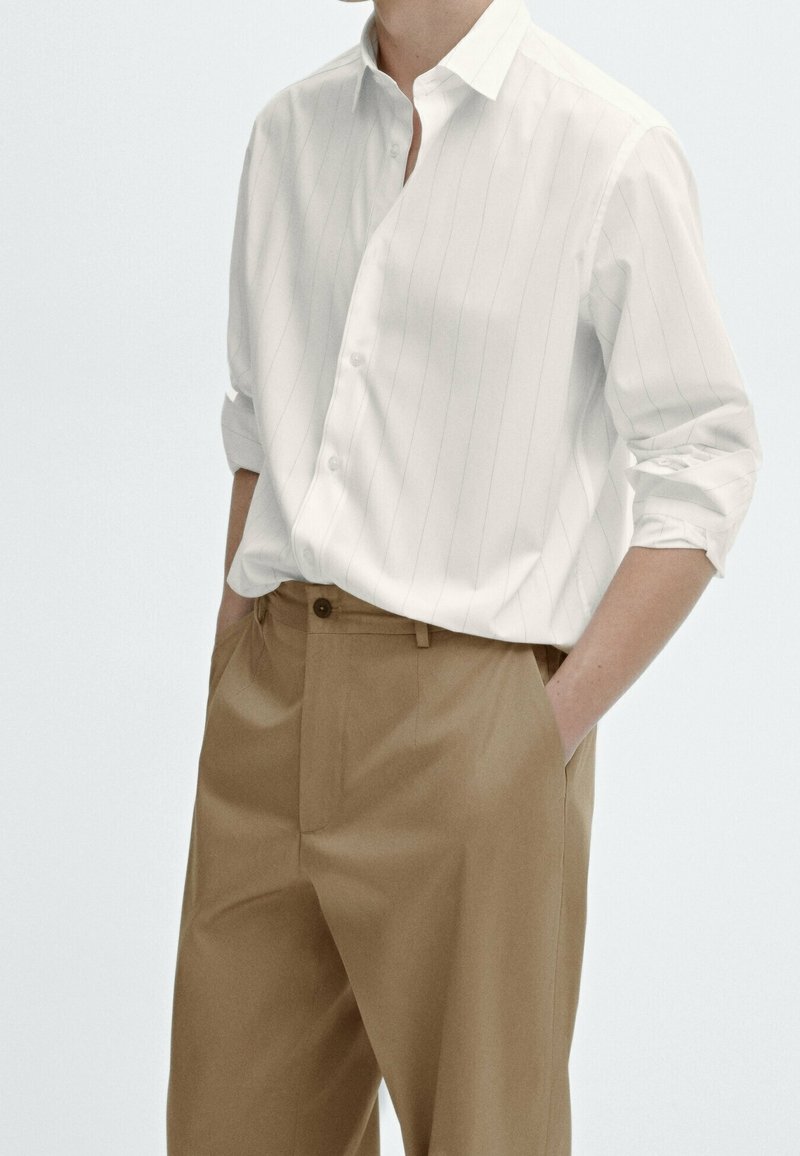 Personne portant une chemise blanche à fines rayures avec manches longues retroussées, rentrée dans un pantalon marron taille haute, mains dans les poches.