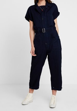 Kobieta ubrana w granatowy kombinezon z krótkim rękawem, z paskiem i kieszeniami z przodu, stojąca na tle białego tła, w białych trampkach wysokich.