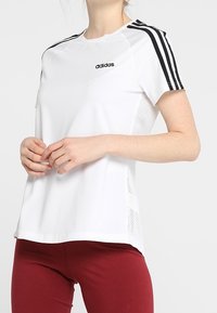 Vit sportt-shirt med svart Adidas-logotyp, med svarta randiga ärmar och nätpaneler vid sidorna. Matcher med röda leggings.