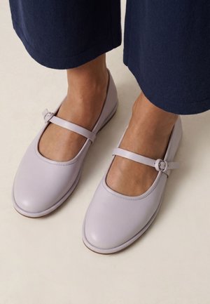 Füße, die hellviolette Mary Jane Schuhe mit Knöchelriemen und marineblaue kurze Hosen auf hellem Hintergrund tragen.