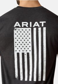 Camiseta gris oscuro con un gráfico de la bandera estadounidense blanca y desgastada en la parte trasera. La palabra "ARIAT" está impresa en letras negritas encima del diseño.