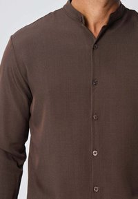 Chemise à manches longues marron en tissu lisse, dotée d'un col mandarin, d'une fermeture à boutons sur le devant et d'un léger éclat.