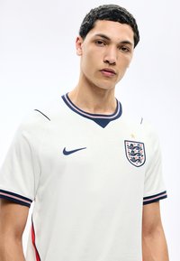 Jonge man die een wit Engeland voetbalshirt draagt met marineblauwe en rode accenten, Nike-logo en Three Lions-embleem op een effen lichte achtergrond.