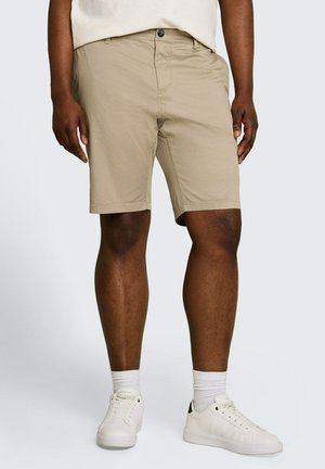 Mężczyzna ubrany w beżowe szorty do kolan, białe skarpety i białe trampki, stojący na jednolitym jasnym tle.