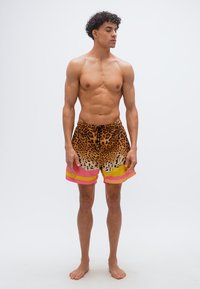 Νεαρός άνδρας με κατσαρά μαλλιά και γυμνό πάνω μέρος του σώματος, φοράει μαγιό λεοπάρ με ροζ και κίτρινες χρωματικές μπλοκ, στέκεται ξυπόλητος σε λευκό φόντο.