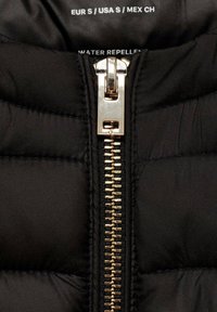 Materiał czarnej pikowanej kurtki puchowej z charakterystycznym złotym zamkiem błyskawicznym. Wnętrze oznaczone rozmiarem i informacją o wodoodporności.