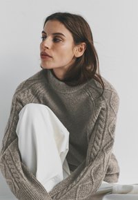 Trui met kabelgebreid patroon in lichtgrijze wol, voorzien van een hoge kraag en oversized mouwen. Witte spijkerbroek met omgevouwen knieën. Minimalistische achtergrond.