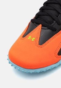 Bottine synthétique orange avec des accents en cuir noir, présentant une surface texturée, des lacets noirs plats et une semelle extérieure à pointes bleu clair. Détail du logo vert.