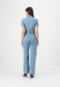 Svetlo modra denim kombinezon s kratkimi zvitimi rokavi, prilegajočim pasom, širokimi hlačami in pasom na pasu. Gladka tekstura, minimalni šivi.