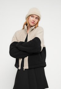 Νεαρή γυναίκα με μπεζ μάλλινο καπέλο και μπεζ-μαύρο τζάκετ sherpa, με τα χέρια σταυρωμένα, κοιτάζει πλάγια σε λευκό φόντο.