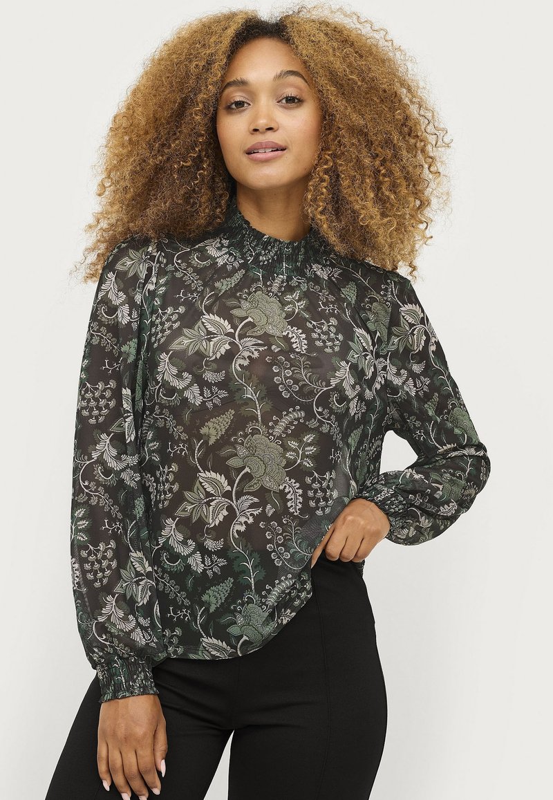 Transparante zwarte blouse met een groen bloemmotief. Heeft een hoge elastische halslijn en lange mouwen met gerimpelde manchetten. Geschreven bij zwarte onderkleding.