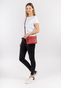 Borsa a tracolla rossa in pelle con due scomparti con zip, texture liscia e tracolla regolabile, indossata da un modello in abbigliamento casual.