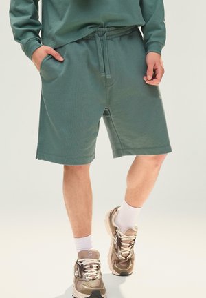 Persoon draagt groene shorts met trekkoord, witte sokken en beige hardloopschoenen, staand met één hand in de zak.