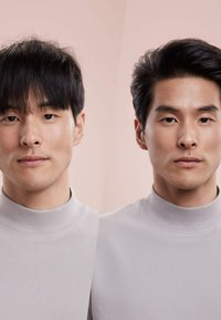 Zwei männliche Personen mit kurzen schwarzen Haaren, die hellgraue hochgeschlossene Hemden tragen und mit dem Gesicht nach vorne vor einem sanften rosa Hintergrund stehen.