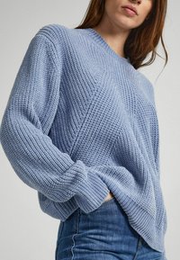 Suéter de punto azul claro con un patrón texturizado, puños acanalados y un corte relajado, combinado con jeans azul oscuro.