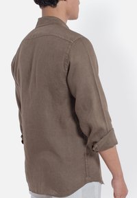 Camicia in lino marrone con vestibilità comoda, maniche lunghe arrotolate e colletto classico. Presenta una cucitura centrale sul retro e una texture sottile.