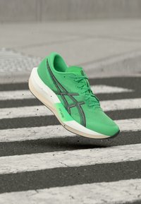 Φωτεινό πράσινο αθλητικό παπούτσι με μαύρο λογότυπο και λευκή σόλα, αιωρούμενο στον αέρα πάνω από διάβαση πεζών με μαύρες και λευκές ρίγες.