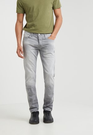 Homme portant un t-shirt vert olive, un jean gris clair usé et des baskets noires, une main dans la poche, sur un fond uni.