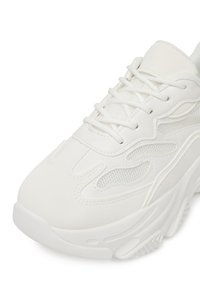 Bílé tenisky se síťovanými a koženými materiály, zakřiveným designem, texturovaným povrchem, výrazným šněrováním a silnou gumovou podrážkou.