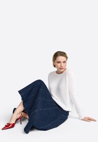 Bela teksturirana jopica, temno modra jeans krila s kontrastnim šivi in rdeče lakirane visoke pete. Modela sedi in gleda naprej.