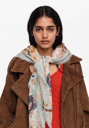 Kobieta z długimi ciemnymi włosami, ubrana w brązową zamszową kurtkę, czerwony sweter i kolorowy, wzorzysty szalik zawiązany wokół szyi.