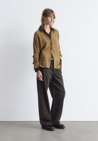 Brown button-up shirt with a darker collar, paired with oversized dark pants and black footwear, set against a minimalistic background.