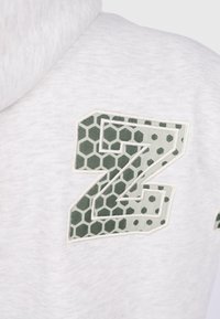 Un hoodie blanc avec un grand patch "Z" décoré de hexagones verts et de points cousu sur la zone de la poitrine.