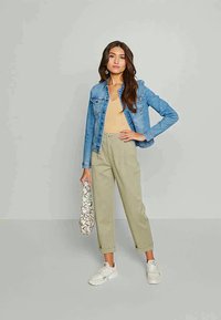 Γυναίκα που φοράει ένα μπλε τζιν μπουφάν, μπεζ αμάνικο μπλουζάκι, κουμπωμένα καμπούρια παντελόνια, λευκά αθλητικά παπούτσια και κρατάει μια λευκή τσάντα με σχέδια.