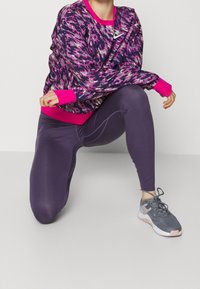 Lila Leggings kombiniert mit einem lebhaften pinkfarbenen und mehrfarbigen sweatshirt mit Muster, das einen runden Halsausschnitt und elastische Bündchen hat. Graue Sportschuhe runden das Outfit ab.