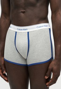Άτομο με σκούρο δέρμα που φοράει γκρι boxer briefs με μπλε λεπτομέρειες και άσπρη μέση με την ετικέτα Calvin Klein, στέκεται μπροστά σε ανοιχτό φόντο.