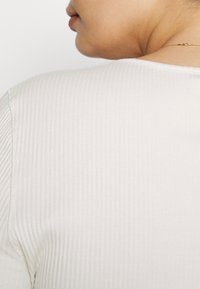 Hladký bílý tričko s žebrovanou strukturou, kulatým výstřihem, viditelnými švy a texturovaným vzorem, zobrazuje zadní část oděvu.