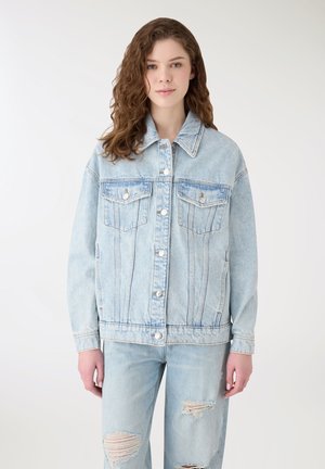 Giovane donna con capelli ricci indossa una giacca di jeans oversize azzurro chiaro e jeans strappati, in piedi davanti a uno sfondo bianco uniforme.