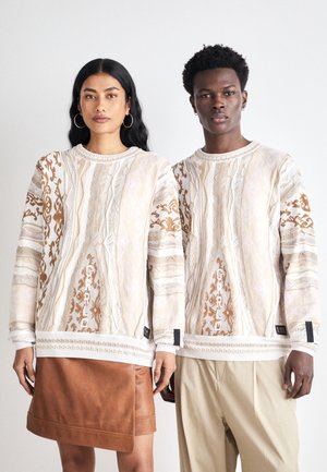 To personer bærer beige og brune mønstrede sweatshirts med strukturerede designs og ribbede manchetter, kombineret med et brunt læderskørt og beige bukser.