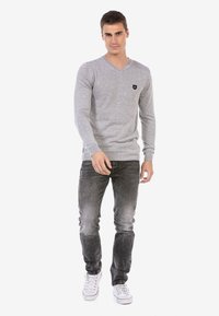 Grauer Langarm-V-Ausschnitt-Pullover mit strukturiertem Finish, mit einem kleinen schwarzen Logo. Getragen mit abgetragenen schwarzen Jeans und weißen Sneakers.