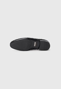 Black flat shoe sole with textured grip and a small rectangular metal brand logo near the arch, photographed on a white background.