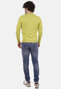 Uomo con capelli ricci, che indossa un maglione verde lime, jeans blu e sneakers bianche, in piedi di spalle su uno sfondo neutro.