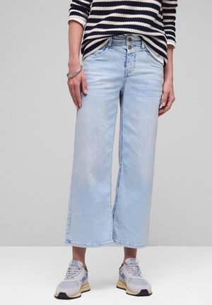 Persoon draagt lichtblauwe wijde cropped jeans, gestreept shirt met lange mouwen en grijze sneakers, staand tegen een neutrale achtergrond.