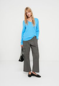 Ανοιχτό μπλε πουλόβερ με λαιμόκοψη σε σχήμα V, γκρι cargo παντελόνι με τσέπες και μαύρα φλατ παπούτσια. Λείες υφές και άνετη εφαρμογή, μινιμαλιστική μαύρη τσάντα.