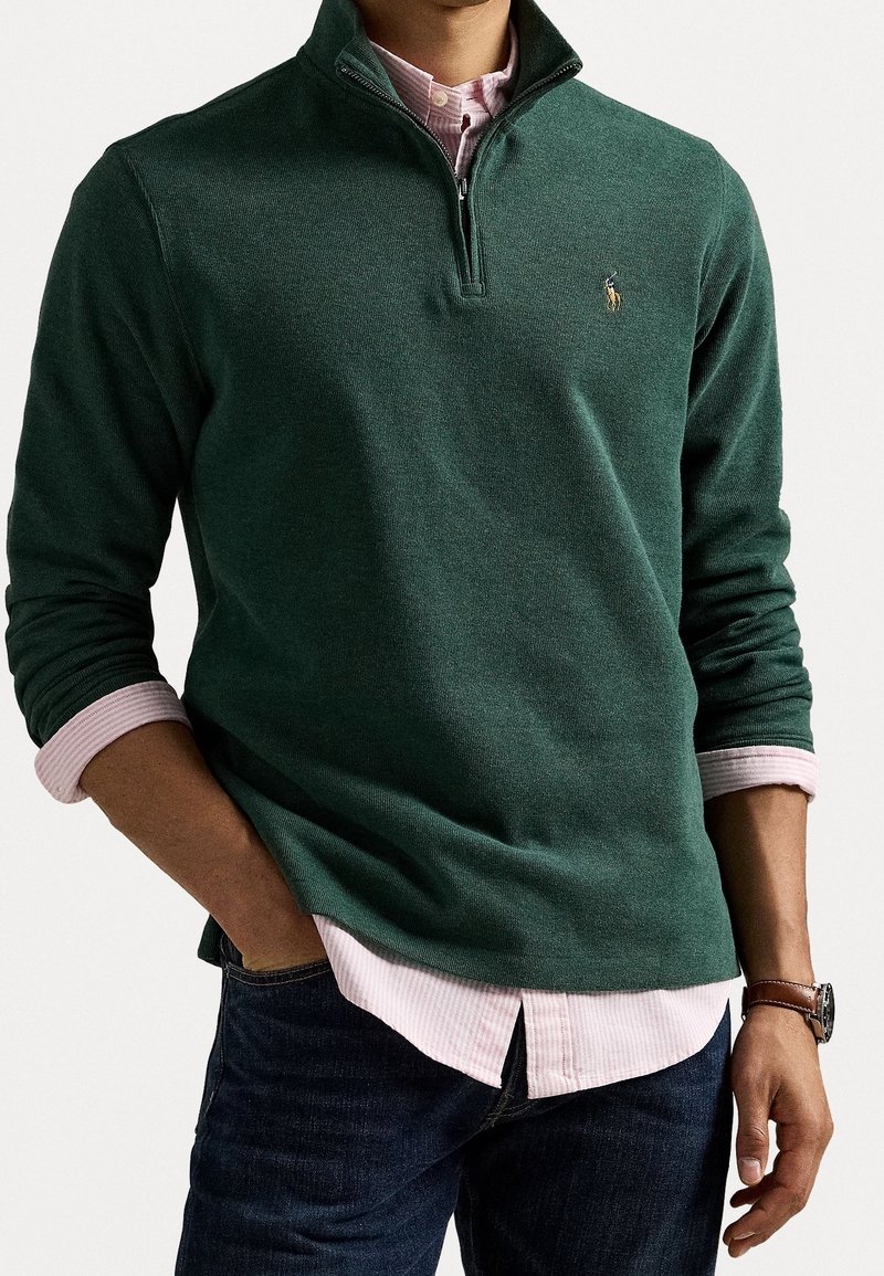 Homme portant un pull vert foncé à demi-fermeture éclair sur une chemise rose clair à boutons, un jean foncé et une montre au poignet gauche, posant avec la main gauche dans la poche.