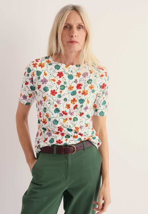 Frau mit langen blonden Haaren, die ein weißes T-Shirt mit Blumenmuster, dunkelgrüne Hosen und einen braunen Gürtel trägt, steht vor einem schlichten Hintergrund.