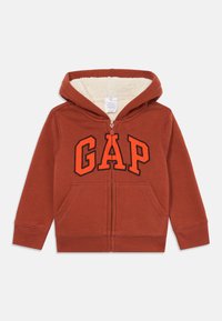 Rød zip-hoodie med kremfarget fleecefôr, med en dristig svart og oransje "GAP"-logo på forsiden og to lommer foran.