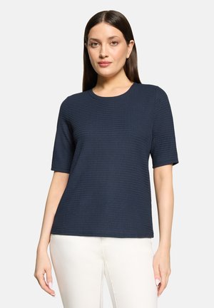 Frau mit glattem dunklem Haar, die ein marineblaues, strukturiertes kurzärmeliges Oberteil und weiße Hose trägt, steht vor einem schlichten hellen Hintergrund.