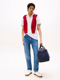 Junger Mann in weißem Poloshirt mit rotem Pullover über den Schultern, blauen Jeans und weißen Sneakers, der eine marineblaue Sporttasche trägt.